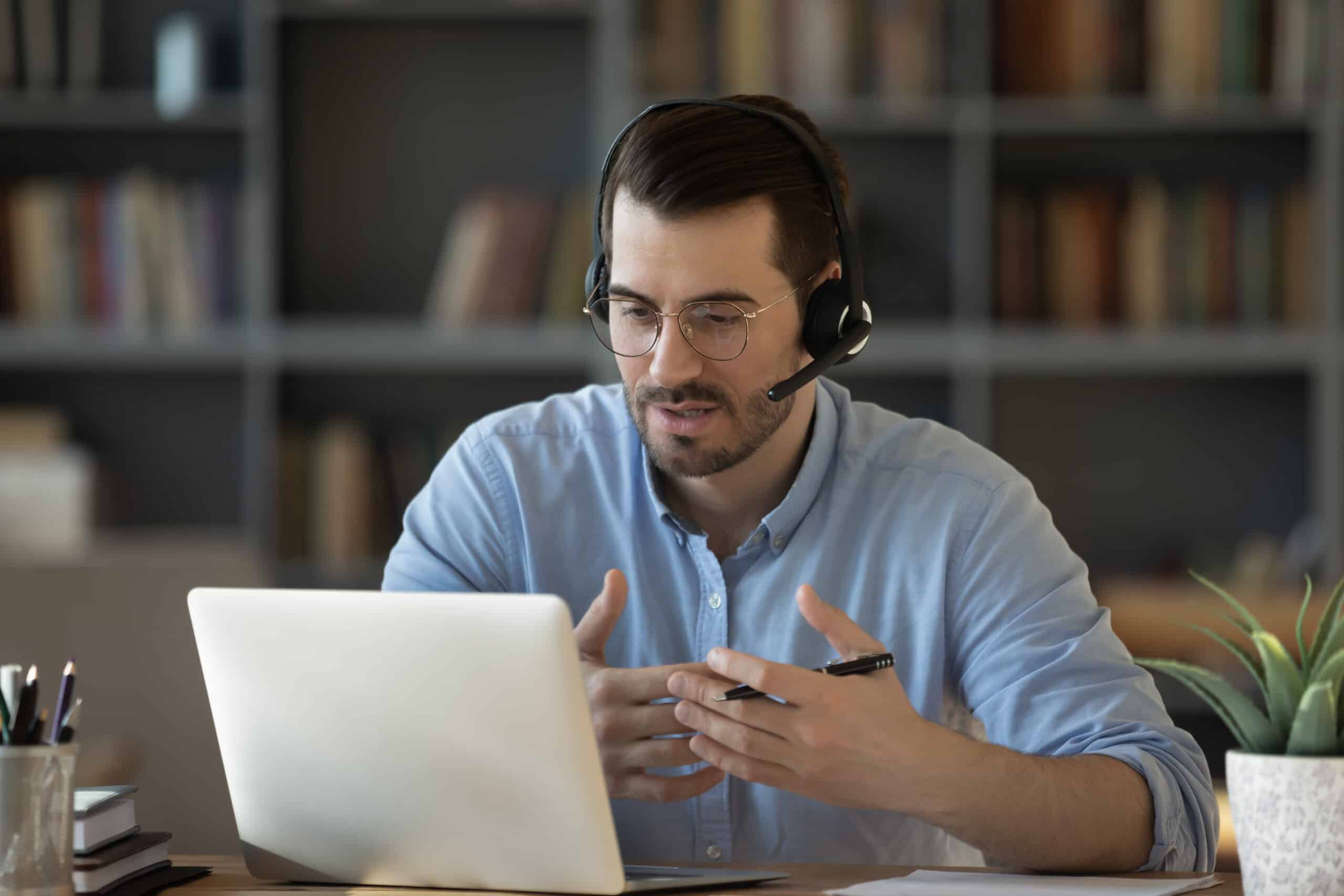 A professional man with dark hair, glasses, and a light blue button-down shirt sits at a wooden desk wearing a black headset with microphone. He is gesturing with both hands while speaking, appearing to be on a video call or virtual meeting. In front of him is an open silver laptop computer.